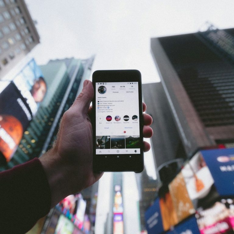 Podcasting Eine Hand hält ein Smartphone, vor großen Werbetafeln in einer Stadtlandschaft.